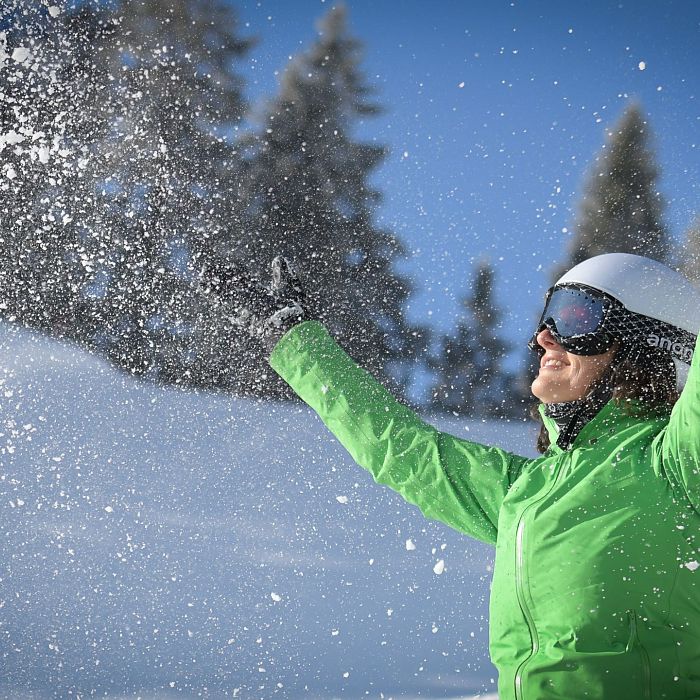 Eine Person in grüner Winterjacke und Skibrille wirft mit Freude Schnee in die Luft. Im Hintergrund sind schneebedeckte Bäume und ein strahlend blauer Himmel.