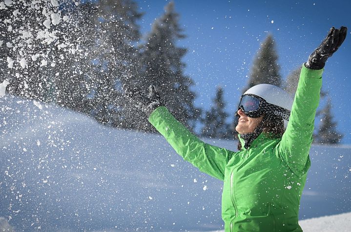 Eine Person in grüner Winterjacke und Skibrille wirft mit Freude Schnee in die Luft. Im Hintergrund sind schneebedeckte Bäume und ein strahlend blauer Himmel.