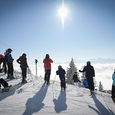 Gruppe von Menschen auf verschneitem Berggipfel bei Sonnenschein, einige mit Skiern, andere in Winterkleidung, umgeben von wolkenverhangenem Tal.