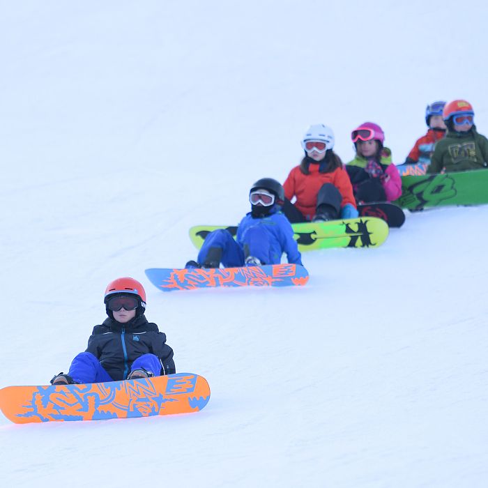 Children in winter gear sit on colorful snowboards on a snowy hill, forming a line. They appear ready for snowboarding under a clear, cold sky.