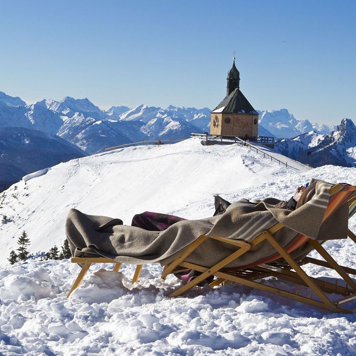 Zwei Liegestühle stehen im Schnee mit Blick auf eine Bergkapelle und eine beeindruckende Alpenkulisse im Hintergrund unter klarem, blauem Himmel.
