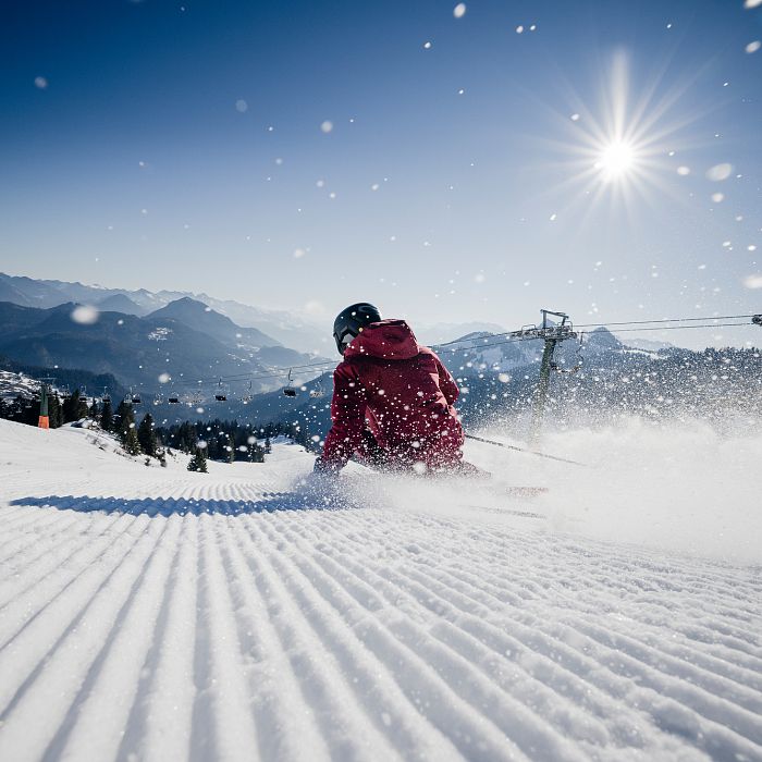 A skier in a red jacket glides down a snowy slope under a bright sun, with mountains in the background and snow sparkling in the air.