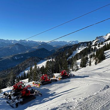 Schneebedeckte Alpenlandschaft mit mehreren Schneeraupen am Hang. Klare blaue Himmel und ferne Bergketten schaffen eine idyllische Winterszene.
