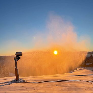 Eine Schneelandschaft bei Sonnenuntergang mit einer Schneekanone im Vordergrund. Der Himmel ist klar und orangefarben, die Sonne steht tief über dem Horizont.