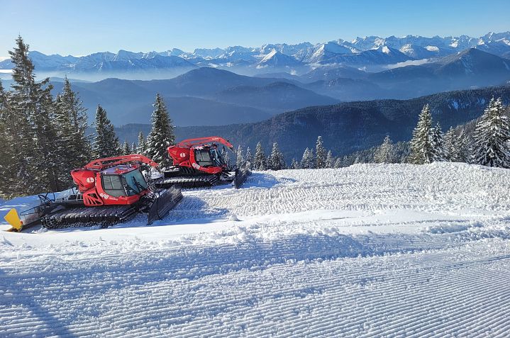 Zwei rote Pistenraupen stehen auf einer schneebedeckten Skipiste mit verschneiten Bergen und Nadelbäumen im Hintergrund unter einem klaren blauen Himmel.