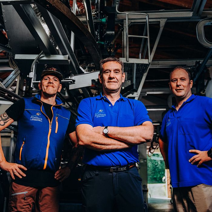 Drei Personen in blauen Polohemden stehen in einer industriellen Umgebung mit Metallstrukturen im Hintergrund und lächeln in die Kamera.