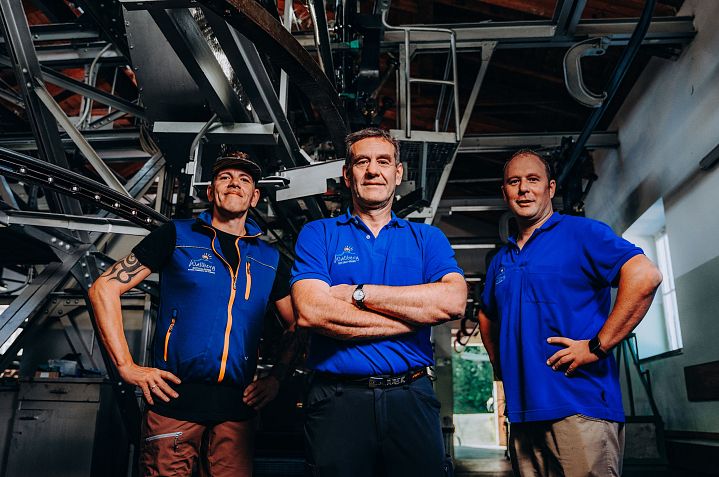 Drei Personen in blauen Polohemden stehen in einer industriellen Umgebung mit Metallstrukturen im Hintergrund und lächeln in die Kamera.