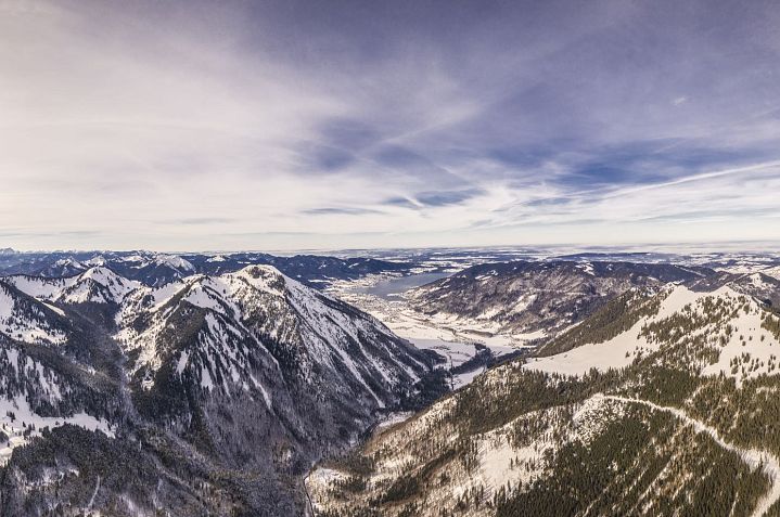 Weite Winterlandschaft mit schneebedeckten Bergen und einem bewölkten Himmel. Im Hintergrund erstreckt sich ein Tal, das von den Bergen umrahmt wird.