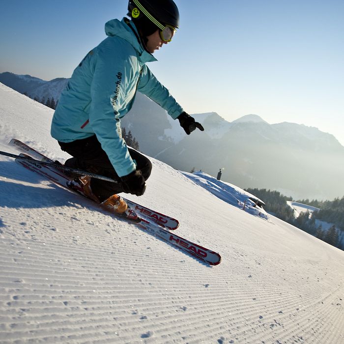 A skier in a blue jacket descends a sunlit snowy slope, surrounded by mountains under a clear sky, with crisp lines in the snow indicating recent grooming.