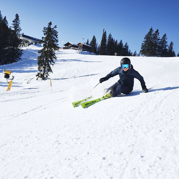 Ein Skifahrer in schwarzer Kleidung fährt dynamisch einen schneebedeckten Hang hinunter. Der Himmel ist klar und blau, umgeben von Tannenbäumen.