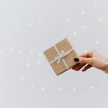 Eine ausgestreckte Hand hält ein kleines, in braunem Papier verpacktes Geschenk mit weißer Schleife vor einem hellen Hintergrund mit sanften Lichtpunkten.