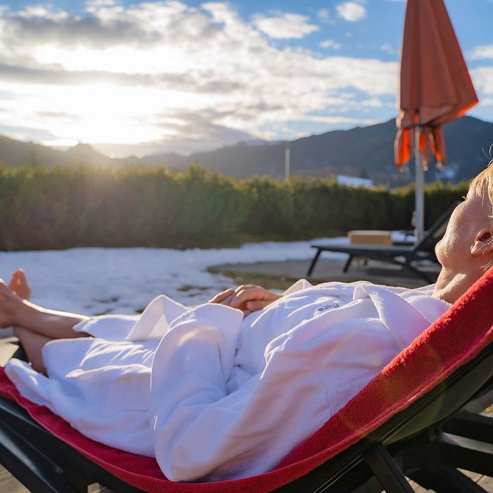 Entspannte Person im Bademantel liegt auf einer Sonnenliege in der Wintersonne, umgeben von Schnee, Sonnenschirm und Berglandschaft im Hintergrund. Komfort und Ruhe.