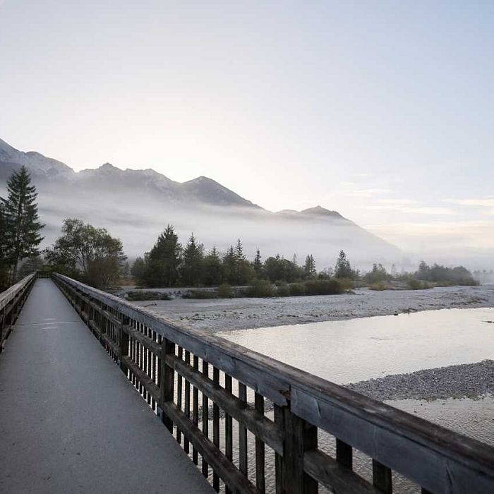 A wooden bridge leads over a river towards misty mountains. Trees and a soft morning sky enhance the tranquil, natural scene.