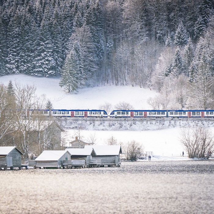 Ein Zug fährt durch eine verschneite Landschaft mit schneebedeckten Bäumen im Hintergrund und mehreren kleinen Holzhütten an einem zugefrorenen Seeufer im Vordergrund.