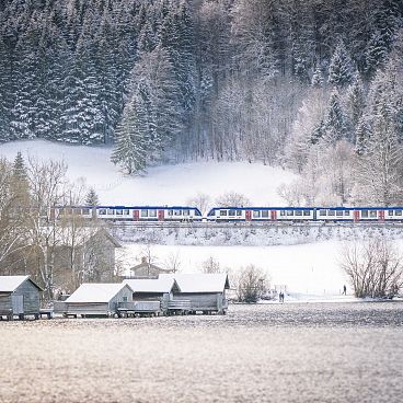 Ein Zug fährt durch eine verschneite Landschaft mit schneebedeckten Bäumen im Hintergrund und mehreren kleinen Holzhütten an einem zugefrorenen Seeufer im Vordergrund.