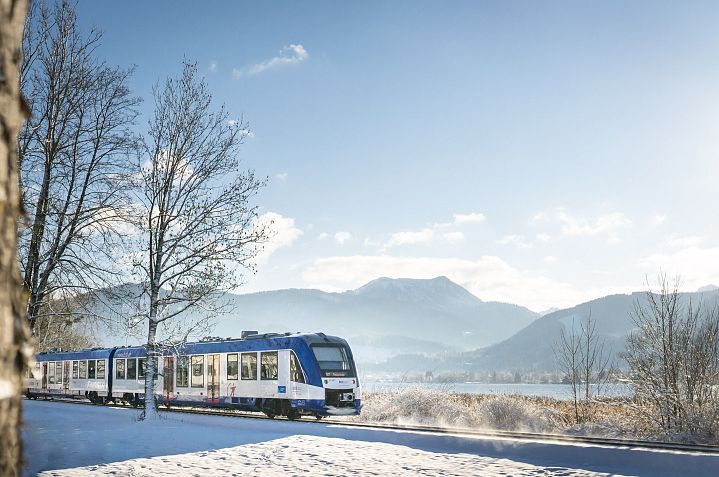 Ein Zug fährt an einem sonnigen Wintertag durch eine verschneite Landschaft mit Bergen im Hintergrund. Die Bäume sind kahl und der Himmel ist klar.