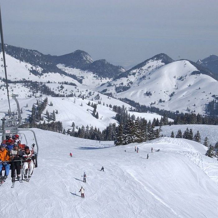 Menschen fahren in einem offenen Sessellift über eine verschneite Winterlandschaft mit Bergen im Hintergrund, während sie Ski und Snowboards tragen.