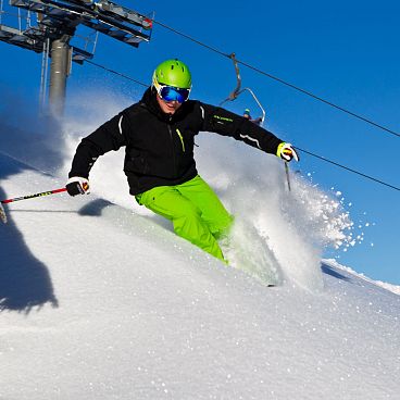 Ein Skifahrer in grüner Hose und schwarzer Jacke fährt dynamisch einen schneebedeckten Berg hinab, während die Sonne auf den klaren blauen Himmel scheint.